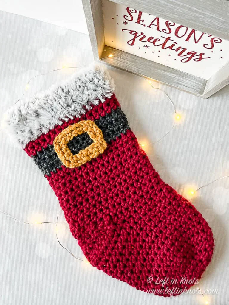 A crocheted stocking made with red bulky yarn and a Santa belt. The stocking has a faux fur trim.