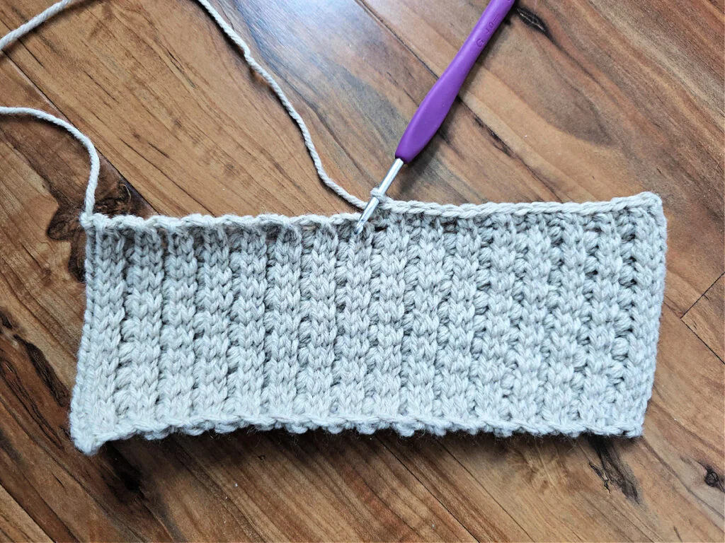 A grey crochet strip with a purple crochet hook against a wooden floor.