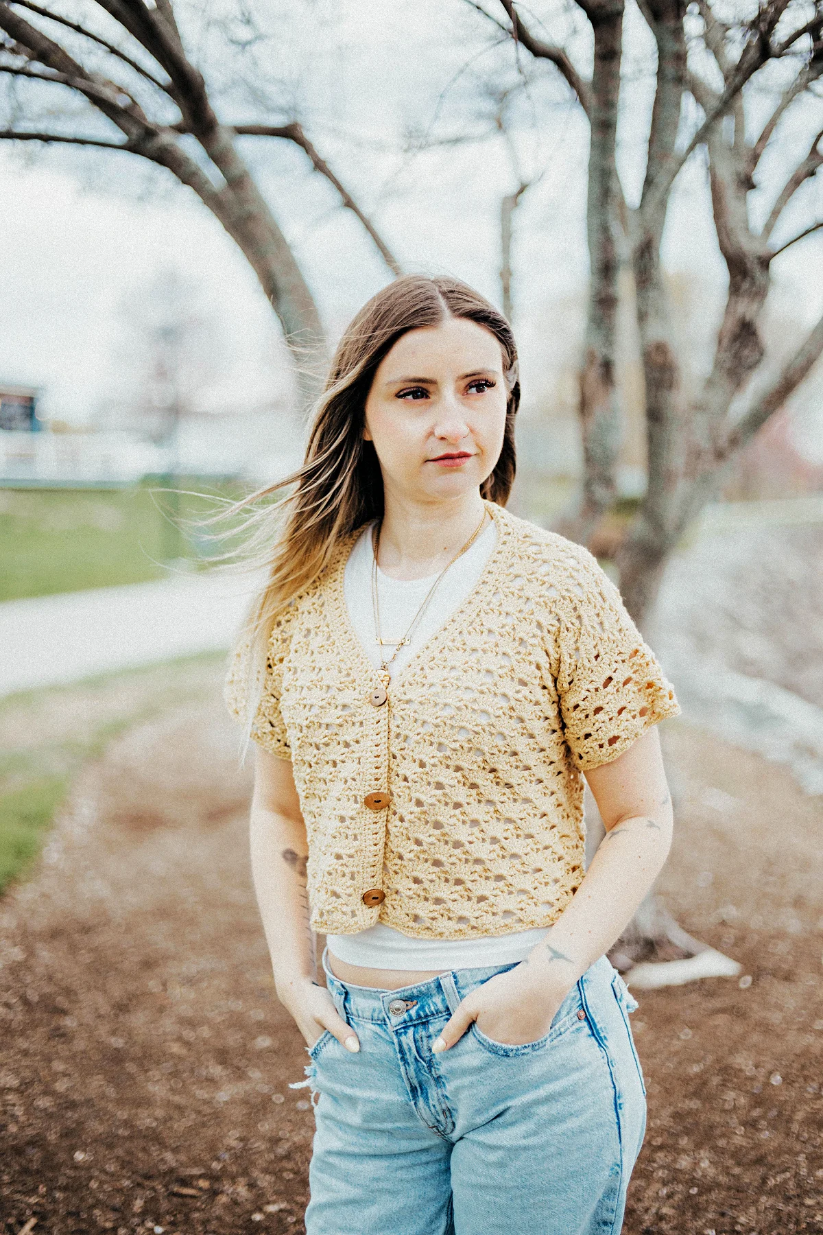 Girl wearing a beige colored cropped crochet cardigan and blue jeans standing in a yard.