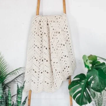 A chunky crochet ripple blanket hanging on a wooden blanket ladder surrounded by plants.