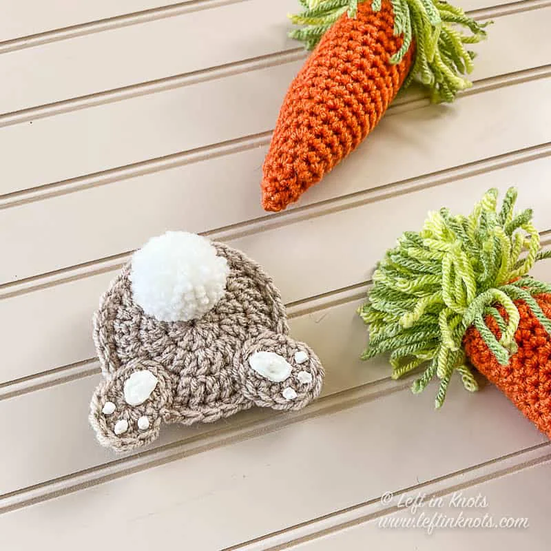 A flat crochet bunny butt applique with a white pom pom tail and hind feet with white paw pads. Displayed next to orange crochet carrots and green grass on a tan background