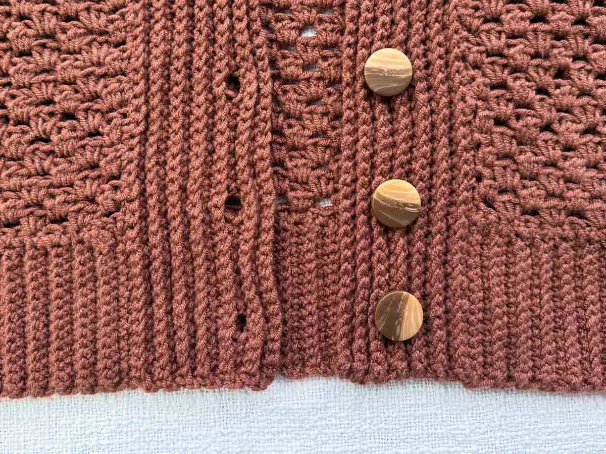 Close-up of the crochet cardigan button band featuring wooden buttons and ribbed edging in "Raisin" Lion Brand Color Theory yarn.