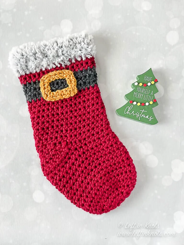A crocheted stocking made with red bulky yarn and a Santa belt. The stocking has a faux fur trim.